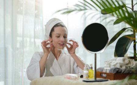 Téléchargez les photos : Beautiful caucasian woman in bathrobe and towel wrapped on head looking at mirror and applying in sheet face mask. - en image libre de droit