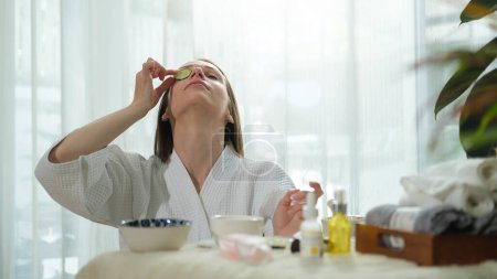 Téléchargez les photos : Jeune femme en peignoir mettant concombre frais sur son oeil tout en faisant un traitement de beauté à la maison. - en image libre de droit