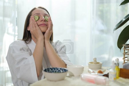 Téléchargez les photos : Beautiful young woman putting slices of cucumber on her eyes and while doing beauty treatment at home.Beauty routine, self care. - en image libre de droit