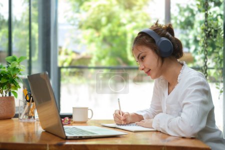Téléchargez les photos : Une jeune femme concentrée portant des écouteurs écrivant des notes, étudiant des cours en ligne via un ordinateur portable. Concept d'enseignement en ligne. - en image libre de droit