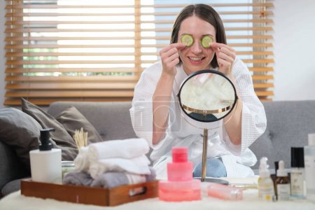 Photo for Young beautiful woman wearing white bathrobe putting slices of cucumber on eyes while doing beauty treatment. - Royalty Free Image