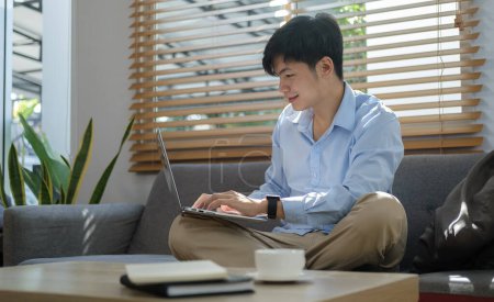 Téléchargez les photos : Smiling asian man sitting on couch and working online on laptop. - en image libre de droit