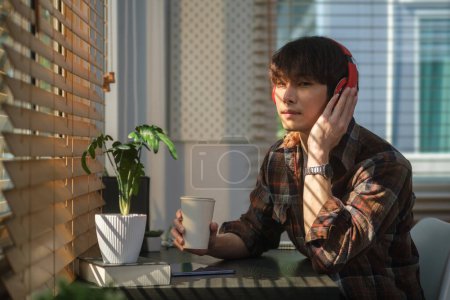 Photo for Carefree young asian man sitting at home office relaxing while listening to music podcast or audio book in head phone. - Royalty Free Image