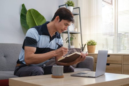 Foto de Tiro de adulto asiático hombre usando un ordenador portátil y tomar notas en el cuaderno mientras se relaja en un sofá en casa. - Imagen libre de derechos