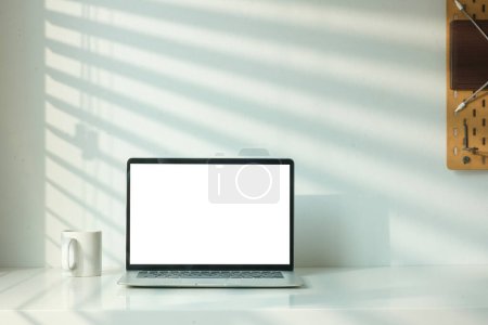 Photo for Front view of laptop computer and coffee cup on table with shadow from the jalousie on a white wall. - Royalty Free Image
