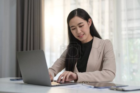Photo for Smiling asian woman executive manager using laptop, communicating online while enjoying work in office. - Royalty Free Image