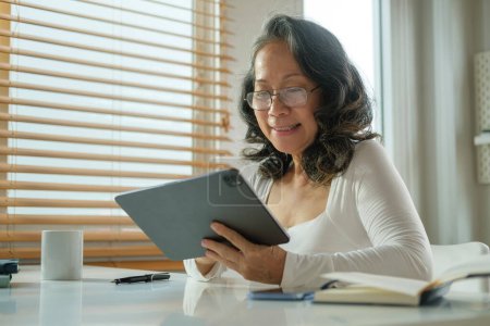 Foto de Mujer mayor feliz con gafas disfrutar de la navegación por Internet en la tableta digital en el hogar acogedor. - Imagen libre de derechos