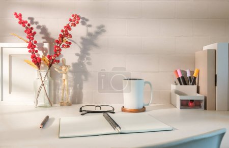 Foto de Cómodo espacio de trabajo con bloc de notas en espiral abierto, vasos y taza de café sobre mesa blanca. - Imagen libre de derechos