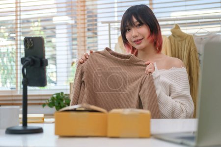 Photo for Pretty Asian female fashion vlogger presenting and showing clothes in front of smart phone, selling online live streaming. - Royalty Free Image
