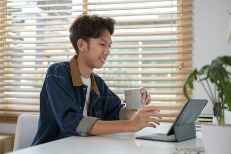 Foto de Feliz asiático freelancer masculino bebiendo café y trabajando en línea en la tableta digital en casa. - Imagen libre de derechos