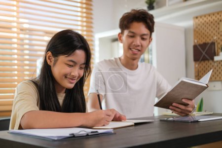 Photo for Smiling asian male tutor assisting teenage girl doing homework  on table at home. - Royalty Free Image