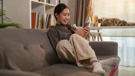 Téléchargez les photos : Souriant jeune femme assise sur le canapé à la maison shopping ou bavarder en ligne sur les réseaux sociaux sur le téléphone mobile. - en image libre de droit
