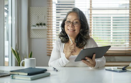 Photo for Happy middle aged asian woman enjoying browsing social media on digital tablet at home. - Royalty Free Image