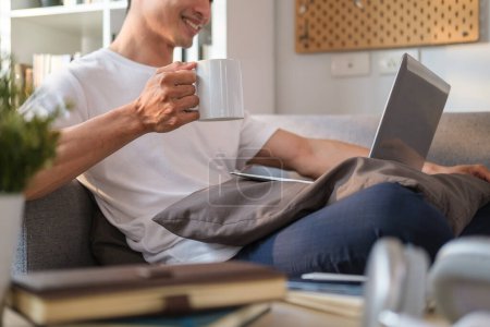 Téléchargez les photos : Homme souriant en vêtements décontractés main tenant tasse de café ad à l'aide d'un ordinateur portable sur le canapé. - en image libre de droit