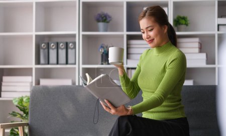 Foto de Mujer joven feliz vistiendo ropa casual bebiendo café y leyendo libro en sofá gris en casa. - Imagen libre de derechos