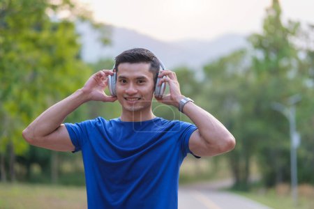 Téléchargez les photos : Homme asiatique athlète en vêtements de sport à l'écoute de la musique sur casque, s 'exercer à l'extérieur au parc. Sport et mode de vie sain. - en image libre de droit