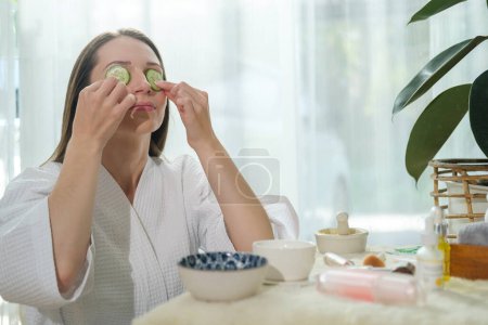 Téléchargez les photos : Young woman in bathrobe putting fresh cucumber on her eye while doing beauty treatment at home. - en image libre de droit