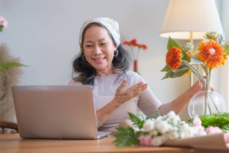 Foto de Mujer mayor sonriente con ramo de flores frescas hablando a través de videollamada en el portátil. Estilo de vida y tecnología de las personas mayores. - Imagen libre de derechos