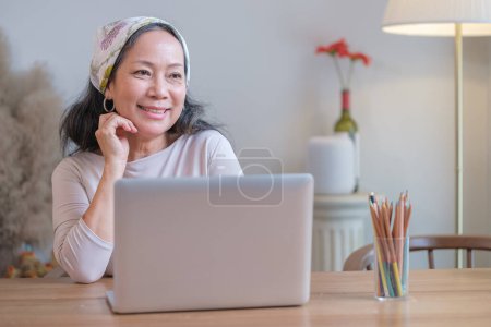 Téléchargez les photos : Femme âgée positive en vêtements élégants à l'aide d'un ordinateur portable à la maison. Objet : - en image libre de droit