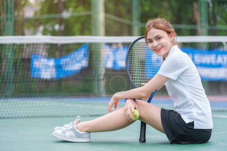 Photo for Female tennis player in sports skirt sitting by the net on court, holding a rocket and smiling to camera. - Royalty Free Image