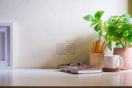 Lieu de travail confortable avec tasse de café, verres, cadre photo et plante d'intérieur sur table blanche.