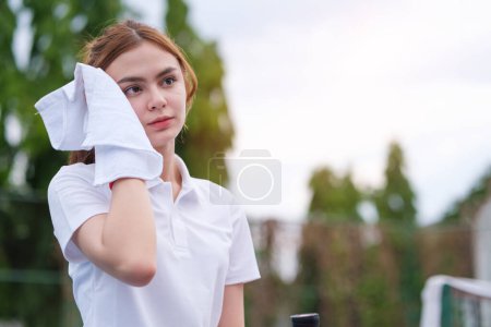 Photo for Tired female tennis player  resting during training session, wiping her face with towel while standing on tennis court. - Royalty Free Image