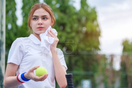 Photo for Beautiful female tennis player  resting during training, wiping sweat with towel while standing on tennis court. - Royalty Free Image