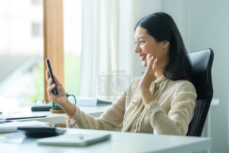Photo for Cheerful millennial Asian woman sitting in comfortable workplace and making video call on mobile phone. - Royalty Free Image