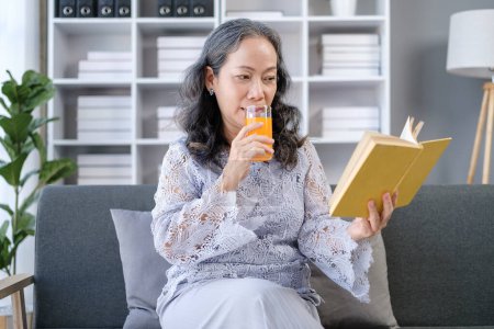 Foto de Mujer jubilada feliz sentada en un cómodo sofá y leyendo un libro interesante, disfrutando del tiempo libre en casa. - Imagen libre de derechos