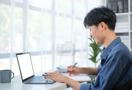 Téléchargez les photos : Satisfait jeune homme d'affaires écrivant sa stratégie sur papier et utilisant un ordinateur portable au bureau. - en image libre de droit