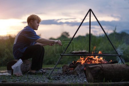 Téléchargez les photos : Homme cuisinant sur un feu de camp pendant un beau coucher de soleil. Concept camping, voyage et loisirs. - en image libre de droit