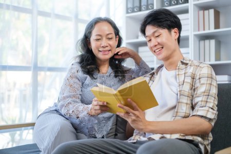 Téléchargez les photos : Femme mûre souriante et jeune fils adulte lisant le livre sur le canapé, passant du temps libre ensemble à la maison. - en image libre de droit
