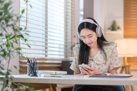 Téléchargez les photos : Jeune femme asiatique écouter de la musique avec casque sans fil et en utilisant un téléphone intelligent tout en étant assis dans le bureau à la maison - en image libre de droit