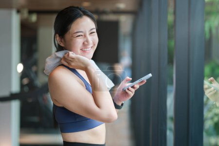 Photo for Delighted sportswoman using smartphone and wiping sweat with towel at gym while relaxing after training. - Royalty Free Image