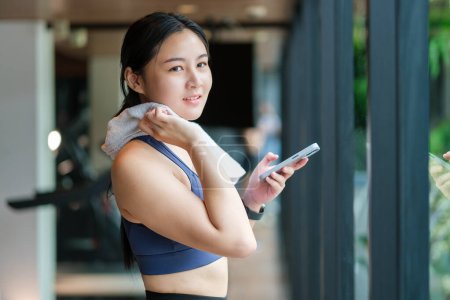 Téléchargez les photos : Femme de fitness souriante essuyant la sueur avec une serviette et utilisant un smartphone dans la salle de gym. - en image libre de droit