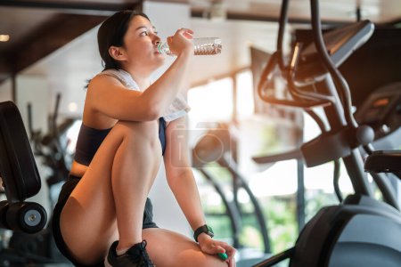 Photo for Attractive fitness woman in sport clothes is drinking mineral water from bottle after workout at gym. - Royalty Free Image