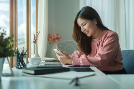 Photo for Smiling female employee sitting in bright office and using smart phone for communicating on social network. - Royalty Free Image