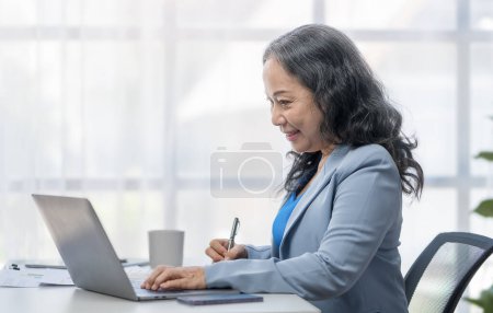 Téléchargez les photos : Femme d'affaires âgée des années 50 souriante utilisant un ordinateur portable et prenant des notes importantes. - en image libre de droit