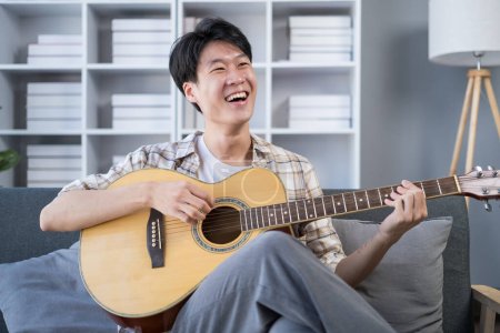Téléchargez les photos : Joyeux jeune homme asiatique chantant et jouant de la guitare sur le canapé dans un salon lumineux et confortable. Loisirs, animations et week-end. - en image libre de droit