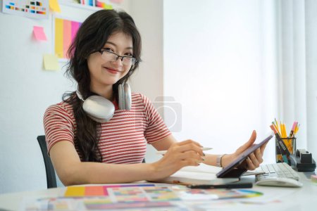 Photo for Cheerful young female designer working on digital tablet and choosing colorful paper charts at desk. - Royalty Free Image