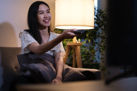 Foto de Relajada mujer asiática sosteniendo control remoto viendo la televisión en la sala de estar por la noche. - Imagen libre de derechos