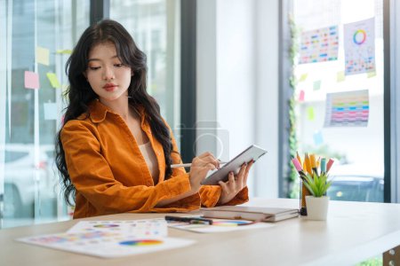 Foto de Joven diseñadora asiática sosteniendo tableta digital y mirando muestras de color, trabajando en el proyecto de diseño en la oficina. - Imagen libre de derechos