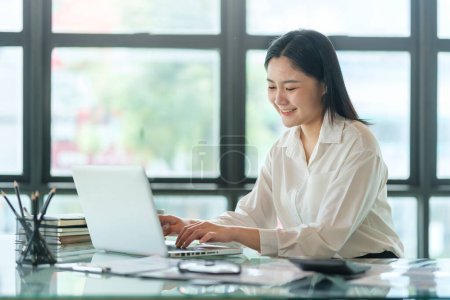 Photo for Young asian female office worker sitting in bright office and working on computer laptop. - Royalty Free Image