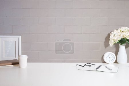 Photo for Home office interior. Picture frame, coffee cup, notebook and pencil holder on white table against brick wall. Copy space. - Royalty Free Image