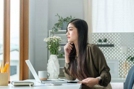 Foto de Pensativo asiático mujer de negocios sentado delante de la tableta en brillante oficina y mirando a través de la ventana. - Imagen libre de derechos