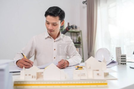 Foto de Guapo arquitecto masculino trabajando con planos, examinando el modelo de proyecto de construcción en el escritorio. - Imagen libre de derechos