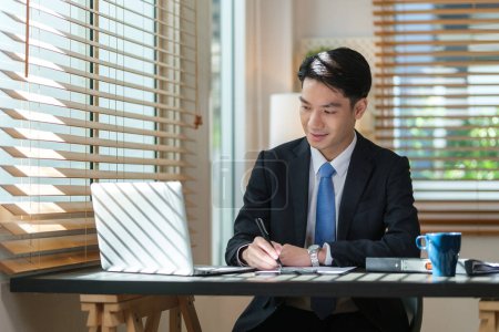 Téléchargez les photos : Focalisé asiatique homme d'affaires écrit des informations importantes sur le bloc-notes et en utilisant un ordinateur portable au bureau de travail. - en image libre de droit