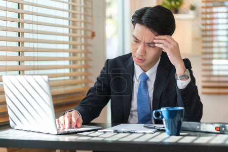 Foto de Joven empresario estresado que sufre de dolor de cabeza sentado en el escritorio de la oficina usando el ordenador portátil. - Imagen libre de derechos