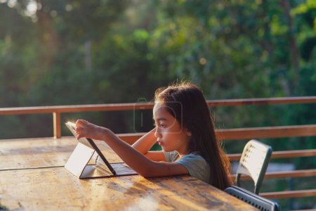 Téléchargez les photos : Petite fille utilisant tablette pad tout en étant assis en plein air pendant le lever du soleil. - en image libre de droit
