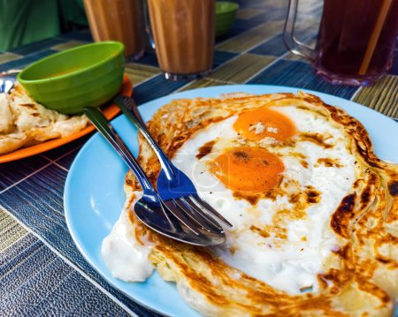 Roti sarang burung, un pratha parece nido de aves con medio cocinar huevos en el medio servido con teh tarik como desayuno básico en Malasia.